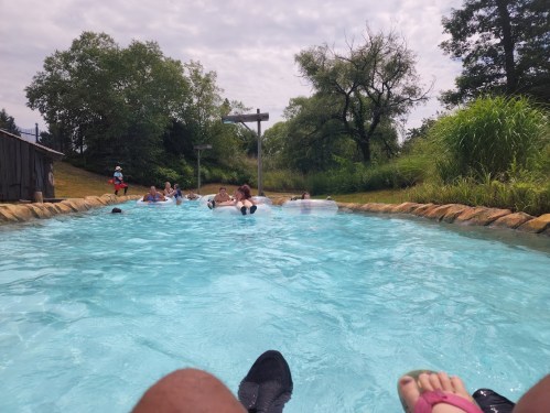 A lazy river with people floating on tubes, surrounded by greenery and a cloudy sky. Feet visible in the foreground.