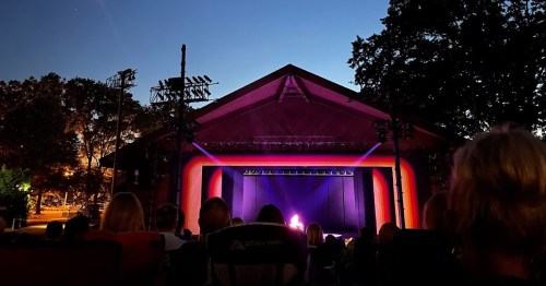 An outdoor theater at dusk, with an audience seated and a performer on stage illuminated by colorful lights.