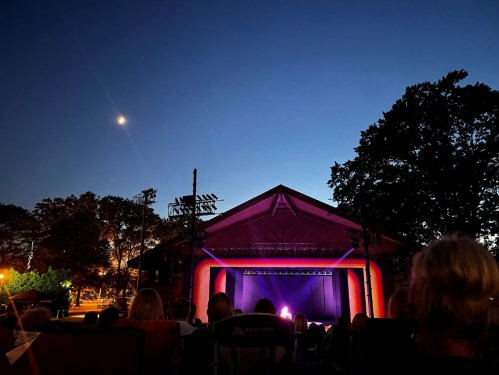 Audience seated in front of a stage at dusk, with a crescent moon visible in the sky above.