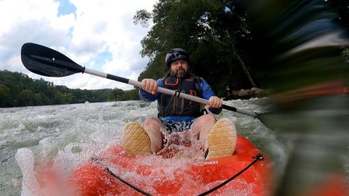 A person paddles a red kayak through turbulent water, wearing a helmet and life jacket, with trees in the background.