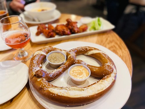 A large soft pretzel with two dipping sauces, accompanied by a plate of chicken wings in the background.