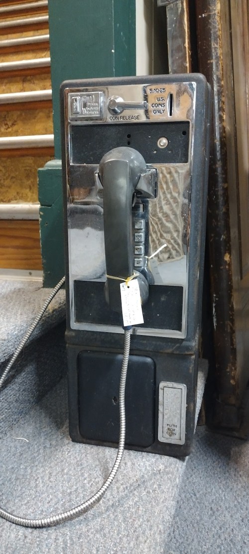 A vintage payphone with a black handset, mounted on a wall, next to a staircase, with a price tag attached.
