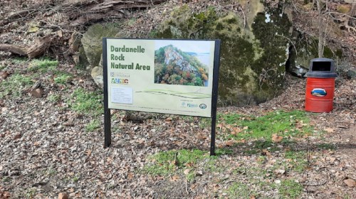 Sign for Dardanelle Rock Natural Area beside a trash can, surrounded by trees and rocky terrain.