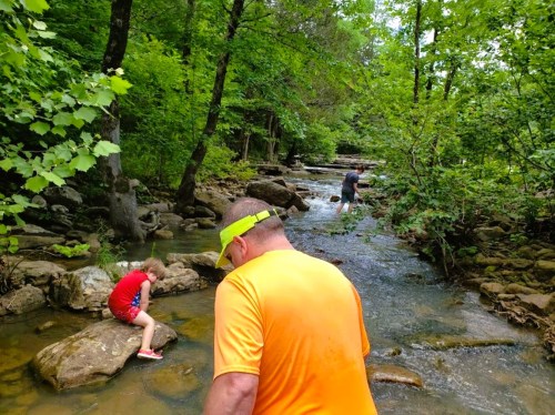 A man in an orange shirt walks along a creek, while a child sits on a rock and another man wades through the water.