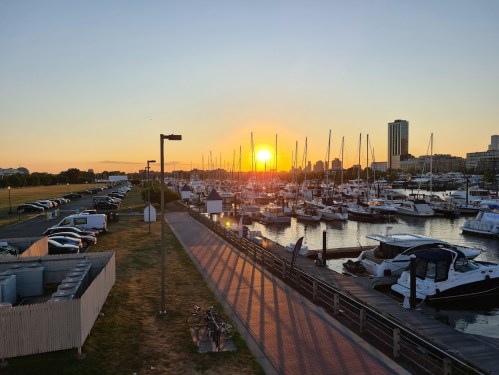 Sunset over a marina filled with boats, with a parking lot and city skyline in the background.