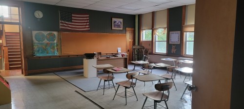 A vintage classroom with wooden desks, a chalkboard, maps on the wall, and an American flag. Natural light from windows.