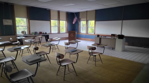 A classroom with empty desks, a podium, and a chalkboard, featuring large windows and an American flag.