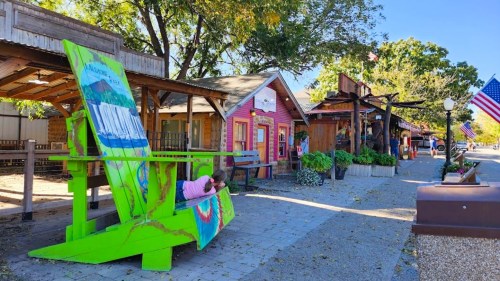 Colorful storefronts line a quaint street, with a large green chair and American flags adding charm to the scene.
