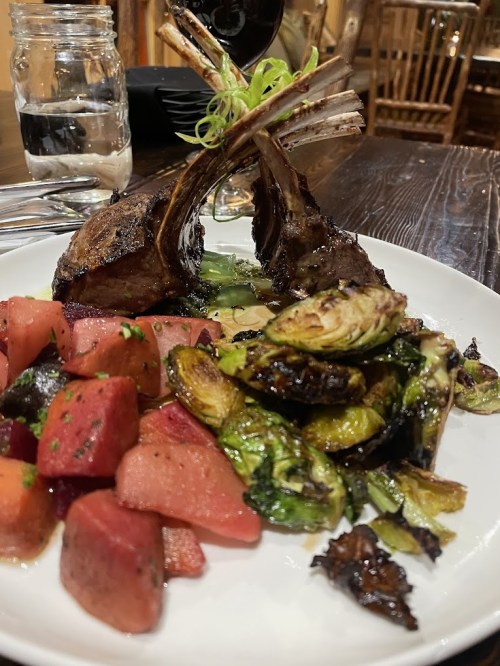 A plate featuring grilled lamb chops, roasted Brussels sprouts, and a colorful watermelon salad, with a glass of red wine.