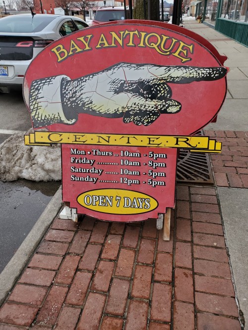 A red and yellow sign for Bay Antique Center, displaying hours and an illustration of a pointing hand. Open 7 days.