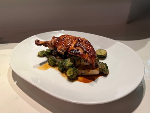 A plated dish featuring roasted chicken leg on a bed of vegetables, garnished with Brussels sprouts and sauce.