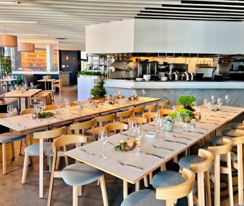 A modern restaurant interior with wooden tables set for dining, featuring greenery and an open kitchen in the background.
