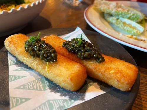 Two golden, crispy croquettes topped with black caviar, served on a black plate with a green logo napkin.