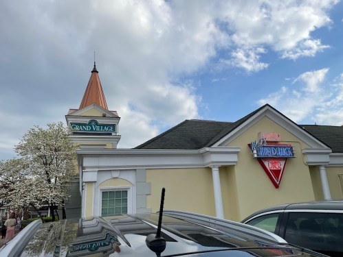 A building with a peaked roof, featuring signs for "Grand Village" and "Waldorf's Waffle House Diner" under a cloudy sky.