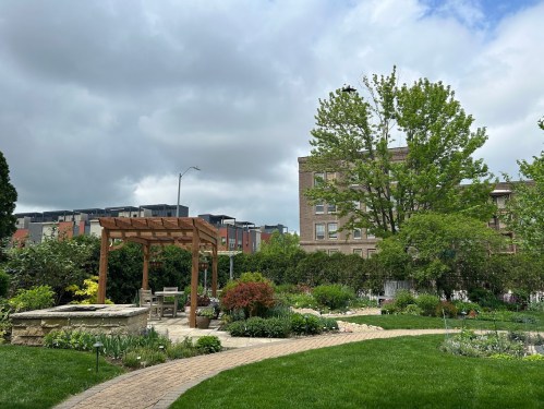 A lush garden with a wooden pergola, green grass, and trees, set against a cloudy sky and urban buildings in the background.