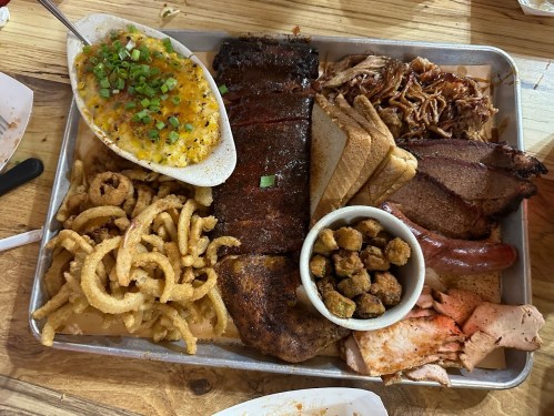 A large platter of barbecue featuring ribs, pulled pork, brisket, sausage, fried okra, and sides of cornbread and onion rings.