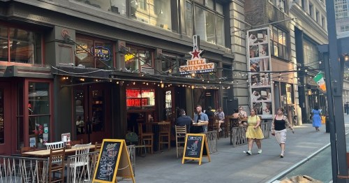 A lively street scene featuring a restaurant with outdoor seating and people walking by in a bustling urban area.