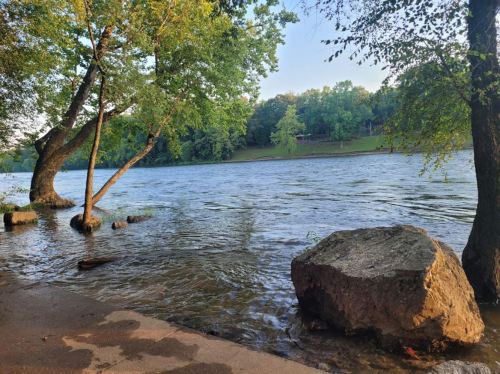 A serene river scene with trees along the bank and a large rock in the foreground, reflecting calm waters.