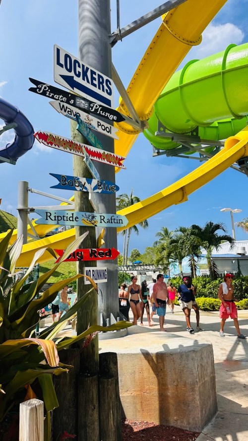 Colorful water park signpost with directions to attractions like "Lockers," "Wave Pool," and various slides. Sunny day.