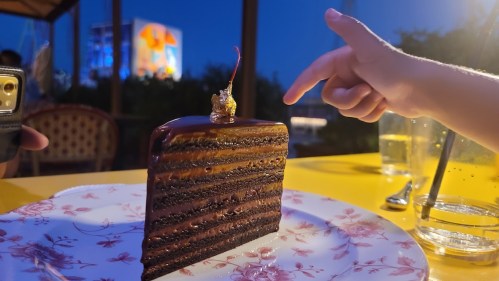 A slice of chocolate cake with a cherry on top, as a child's hand reaches toward it in a dimly lit setting.