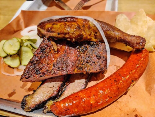 A platter of assorted smoked meats including brisket, sausage, chicken, and pickles, served on brown paper.