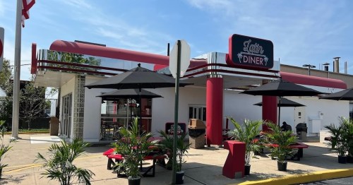A retro-style diner with a red and white exterior, outdoor seating, and umbrellas on a sunny day.