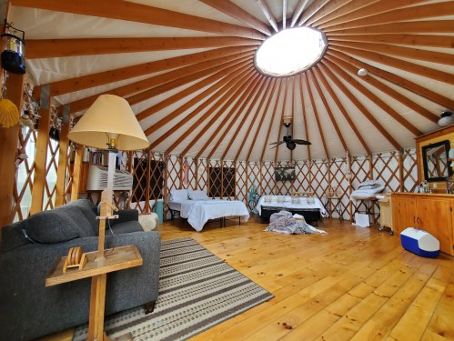 Interior of a yurt with wooden beams, a circular skylight, a sofa, and a cozy, rustic atmosphere.