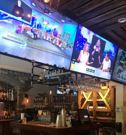 A bar with two large TVs showing talk shows, featuring a lively atmosphere and a well-stocked drink display.