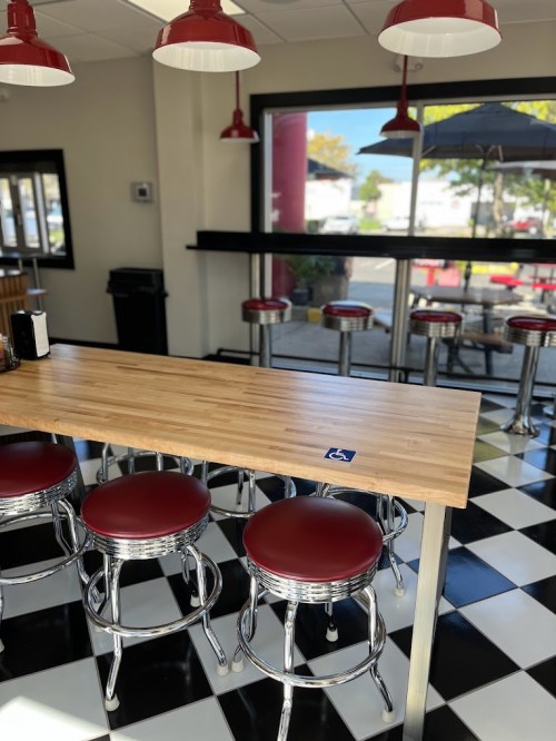 A modern diner interior with a wooden table, red stools, and a black-and-white checkered floor. Large windows provide natural light.