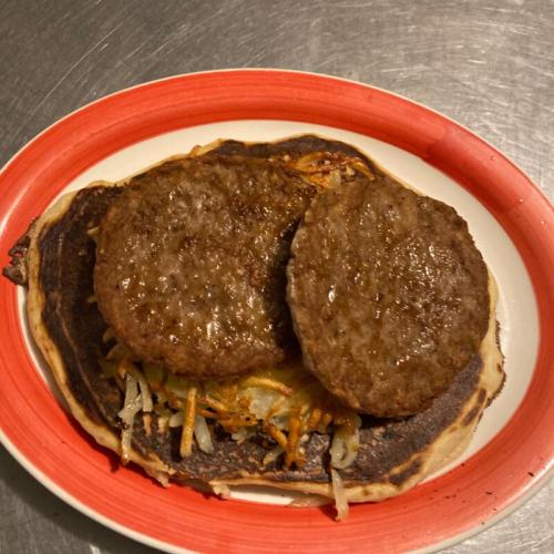 A plate with two cooked sausage patties on a burnt pancake topped with sautéed onions and carrots.