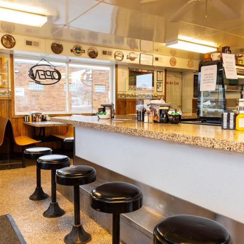 A cozy diner interior featuring a counter with stools, wooden paneling, and a "OPEN" sign in the window.