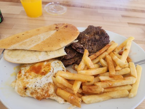 A plate with two pieces of bread, a fried egg, beef, and a serving of French fries, with a drink in the background.
