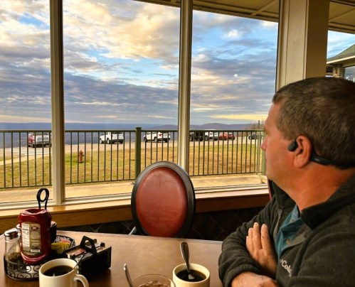 A person sits at a table by a window, gazing at a scenic view of clouds and mountains outside.