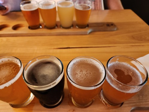 A wooden table with a flight of craft beers in various colors and styles, with more beers in the background.