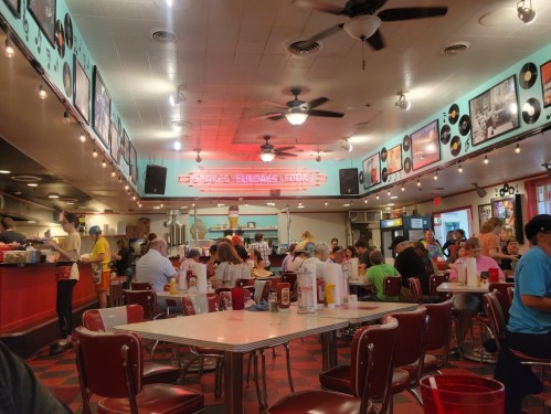 A bustling diner interior with red booths, a checkered floor, and retro decor, filled with patrons enjoying their meals.