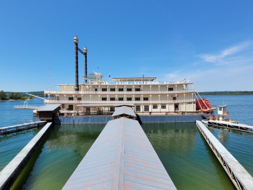 A large riverboat with two smokestacks docked at a pier on a calm lake under a clear blue sky.