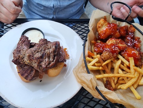 A plate with grilled steak on bread and a basket of crispy chicken wings with fries, drizzled in sauce.