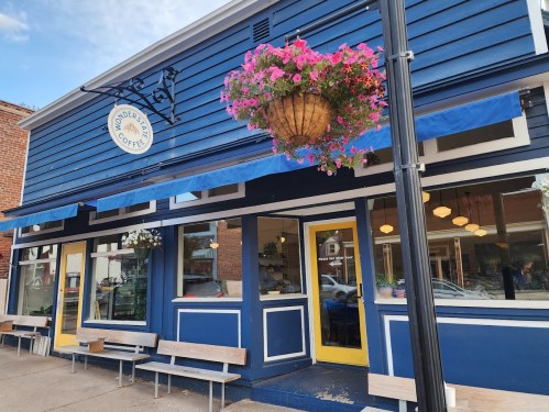 A blue coffee shop with hanging flower baskets and outdoor seating, featuring large windows and a welcoming entrance.