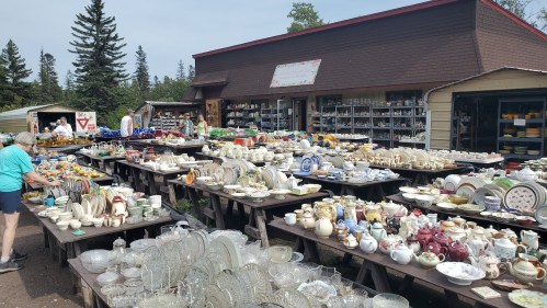 A bustling outdoor market filled with tables of various pottery, glassware, and collectibles under a sunny sky.