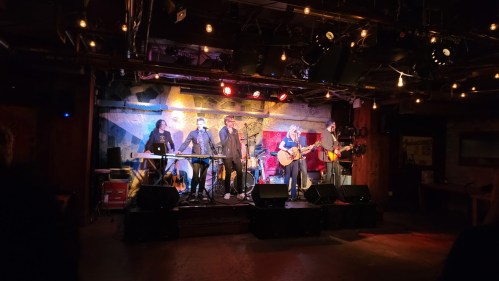 A live band performs on stage with instruments and colorful lights in a cozy venue. Audience members are visible in the foreground.