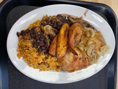 A plate of seasoned chicken with fried plantains, rice, and black beans, served on a black tray.
