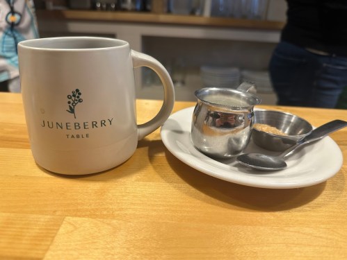 A white mug with "Juneberry Table" logo next to a small creamer, sugar, and a spoon on a plate.