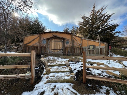 A rustic, round building with a wooden door, surrounded by snow and trees, leading to a stone pathway.