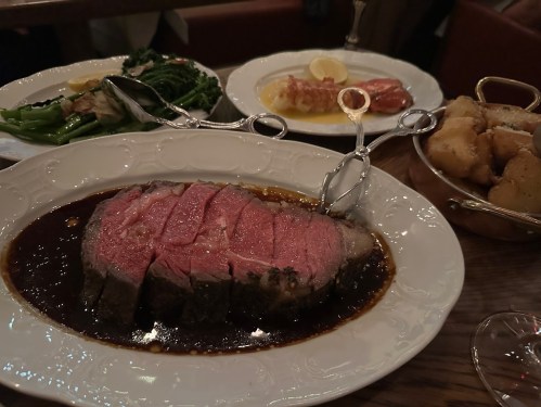 A plate of sliced roast beef in sauce, accompanied by sides of vegetables and seafood in a restaurant setting.