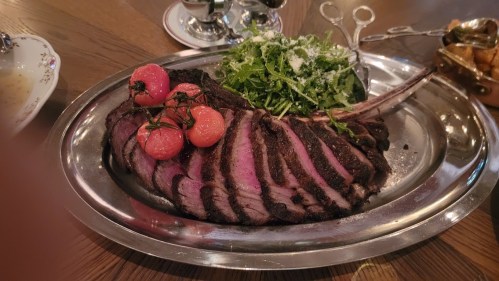 A beautifully cooked ribeye steak on a silver platter, garnished with cherry tomatoes and a side of arugula salad.