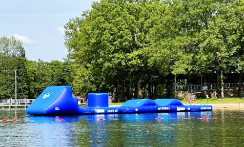 A blue inflatable water obstacle course on a lake, surrounded by trees and a clear sky.