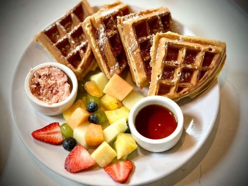 A plate of waffles with fruit, butter, and syrup, featuring strawberries, blueberries, and assorted melons.