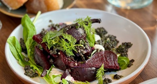 A plate of roasted beets on a bed of greens, topped with capers and fresh herbs, served with a creamy sauce.