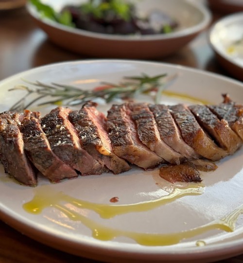 Sliced steak on a decorative plate, drizzled with olive oil, with a side of salad in the background.