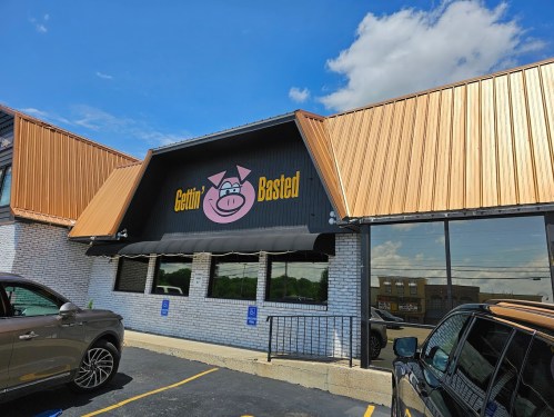 Exterior of "Gettin' Basted" restaurant with a pig logo and a copper-colored roof under a blue sky.